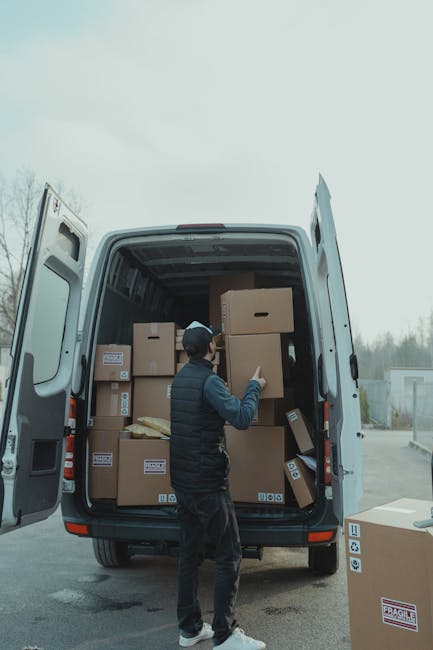 A person wearing a dark vest, black pants, and a cap is standing at the open rear doors of a large white van, which is parked outdoors on a paved surface. Inside the van, numerous cardboard boxes of various sizes are visible, some with printed labels and barcodes, suggesting they are packed for a home relocation or moving service. The individual appears to be placing or adjusting a box near the middle of the van, with additional boxes stacked towards the back and sides, some partially tilted or resting on others. To the side of the van, there is a loose cardboard box with labels indicating fragile contents. The scene is set during daylight with overcast skies, and a background of trees and a distant building suggests a suburban or semi-rural environment. This image illustrates the loading process in furniture transport, typical of professional removals like those offered by Man and Van Pimlico, supporting the theme of packing and moving in a residential or commercial property transition.
