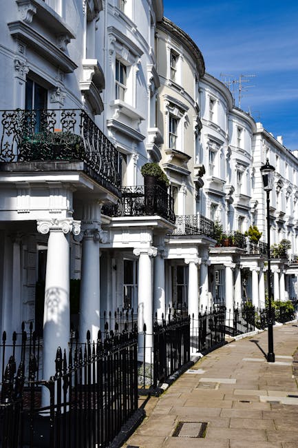 A row of white, Victorian-style terraced houses with ornate architectural details, including decorative cornices and black wrought iron balconies, situated along a paved pavement under a partly cloudy sky. The buildings feature large sash windows and small front gardens enclosed by black metal railings. In the foreground, a man and van service provider, Man and Van Pimlico, is seen loading a variety of household items, including cardboard boxes, a wooden wardrobe, and wrapped furniture, onto a van parked nearby. The items are carefully wrapped in plastic or blankets to prevent damage during transport, and the loading process is taking place outside the property’s front entrance. The scene highlights the logistics of home relocation, with care taken to carry and load furniture and packing materials, supported by the presence of equipment such as trolleys and straps. The environment appears clean and well-organized, with natural daylight illuminating the street scene for a professional packing and moving operation related to end-of-tenancy removals in Pimlico.