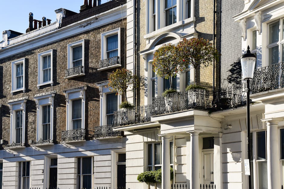 A street view on Bessborough Gardens in Pimlico, showing a residential area with white, multi-storey Victorian-style townhouses featuring decorative cornices and balcony railings. Tall trees with green foliage line both sides of the street, and several parked cars, including a black SUV and a white van, are visible along the curb. The street has traffic lights displaying red signals at intersections, and pedestrians are seen waiting at the crossings. In the foreground, a person is standing near the corner of the sidewalk, possibly reading or holding a device, while another person is walking on the pavement. The sky is overcast with gray clouds, casting diffuse lighting over the scene. This setting could relate to home relocation activities, where furniture and boxes are packed and prepared for transport in a moving process managed by a professional removal service such as Man and Van Pimlico, who specialises in end-of-tenancy and house removals within Pimlico.