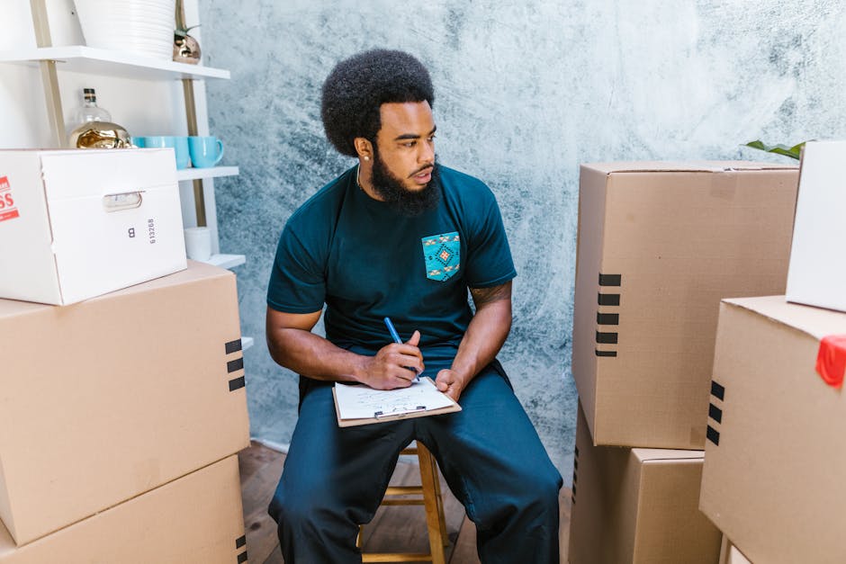 A man with dark curly hair and a beard, wearing a navy blue t-shirt with a logo on the chest, is seated on a wooden stool inside a room with a textured light blue wall. He is holding a pen and a clipb