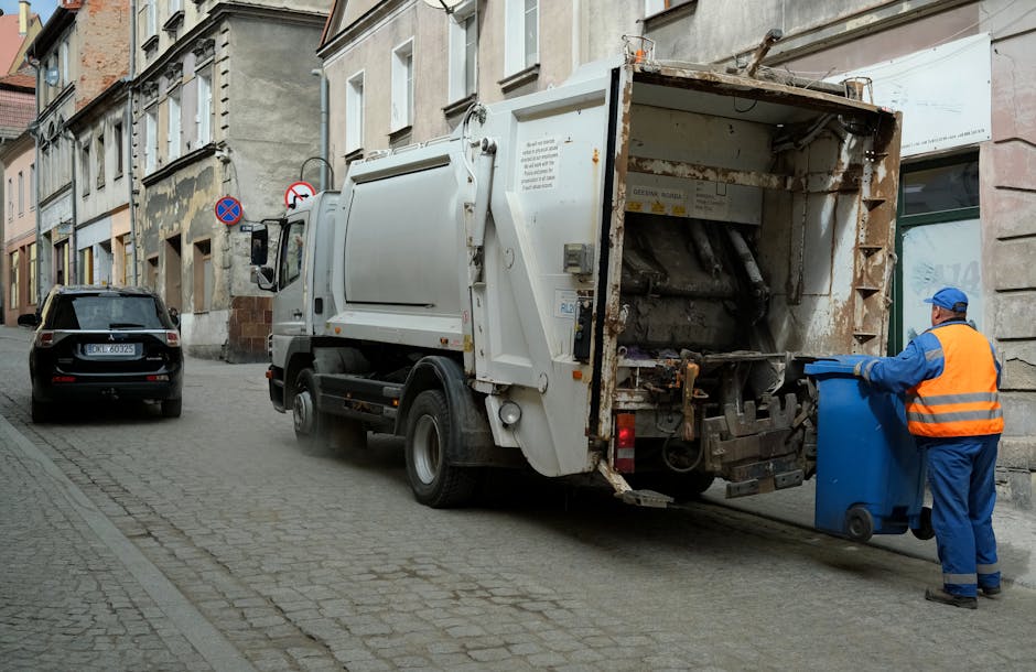 A sanitation worker dressed in a blue uniform and orange safety vest is seen emptying a blue wheeled bin into a white waste collection truck parked on a narrow cobblestone street in Pimlico. The truck's rear compartment is open, revealing mechanical components and collection mechanisms for rubbish. The street is lined with old, weathered buildings with multiple windows and facades showing signs of age and patchwork repairs. A black car is parked further down the street, and traffic signs indicating parking restrictions are visible on the building walls. The scene is outdoors during daylight with natural lighting, illustrating a typical waste collection process that is part of the home or commercial property waste management and clearance services, often linked with relocation or clearance projects carried out by companies like Man and Van Pimlico.