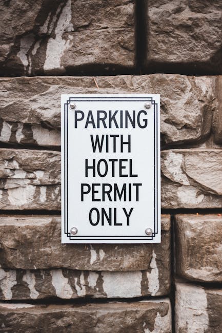 A rectangular metal sign mounted on a rough stone wall, displaying the message 'PARKING WITH HOTEL PERMIT ONLY' in bold black capital letters. The sign is secured with four screws, one at each corner, and is situated in an outdoor urban setting. In the foreground, part of a moving van or vehicle is visible, suggesting a home relocation or furniture transport process. The image reflects the context of loading or unloading activities near a residential or hotel area, relevant to house removals and professional moving services by Man and Van Pimlico.
