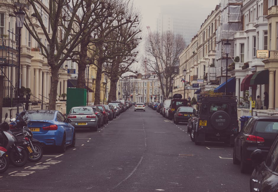 A quiet urban street scene in Pimlico, showing a wide road lined with white Victorian-style terraced houses on both sides, with some featuring rounded bay windows and decorative cornices. Several cars are parked along the curb, including a black SUV, a white van, and a small blue car, with a few vehicles visible further down the street. Mature trees with green foliage flank the road, some extending over the pavement, creating partial shade. Pedestrians are waiting at a pedestrian crossing with traffic lights, which are currently red, and a person in a beige coat and backpack stands near the corner. Visible equipment includes streetlights, and the sky is overcast with gray clouds, indicating typical London weather. This scene depicts a residential area suitable for house removals or furniture transport, and it aligns with services offered by Man and Van Pimlico for home relocation and packing and moving logistics.