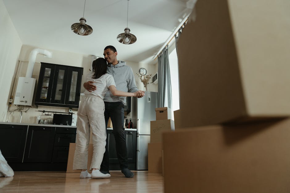 Inside a well-lit apartment during a home relocation, a man and woman are embracing, with the woman standing on a wooden floor and the man slightly taller, both appearing happy. The kitchen area behind them features black cabinetry with glass-fronted upper cabinets, a white boiler mounted on the wall, and a small black countertop with a few items on it. Several cardboard moving boxes of different sizes are stacked on the floor near the window, which has light blue curtains and is partially open, letting in natural daylight. The ceiling has two metallic pendant lights hanging above, casting soft illumination. The scene captures the final moments of packing or a farewell in the early stages of furniture transport and packing for the move, with the presence of a moving company, Man and Van Pimlico, supporting the logistics of this house removal on Lupus Street.