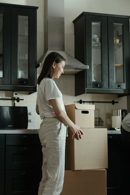 A person wearing a dark t-shirt is standing behind a kitchen counter filled with moving boxes and packing materials, including cardboard boxes, a small potted plant, and packing tape. The scene shows the interior of a home being prepared for a house move, with several cardboard boxes stacked on the floor and on the counter, some partially filled or open. The kitchen features dark cabinetry and a light-colored wall, with the individual appearing to be involved in packing or organising belongings as part of a home relocation process. The image is taken from an adjacent room, looking through a doorway into the kitchen, capturing the loading and packing stage associated with house removals, coordinated by Man and Van Pimlico.