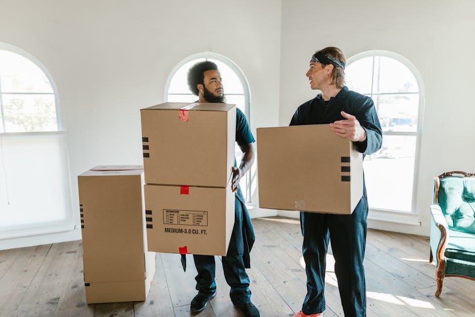 Two professional movers from Man and Van Pimlico are inside a bright, spacious room with white walls and large arched windows, engaged in a home relocation process. One mover, an African man with a beard, is standing next to a stack of three cardboard moving boxes, holding one box with both hands, while the other mover, a Caucasian man wearing a black headband, is gesturing as he holds a single large cardboard box. The boxes are taped and labeled, indicating packing and moving preparations. The room features wooden flooring and minimal furniture, including a green upholstered chair with wooden accents positioned along the wall to the right. The scene captures the loading process, with the movers preparing to transport the boxes, supported by a professional removals service like Man and Van Pimlico, highlighting the logistics involved in furniture transport and household packing for a local move.